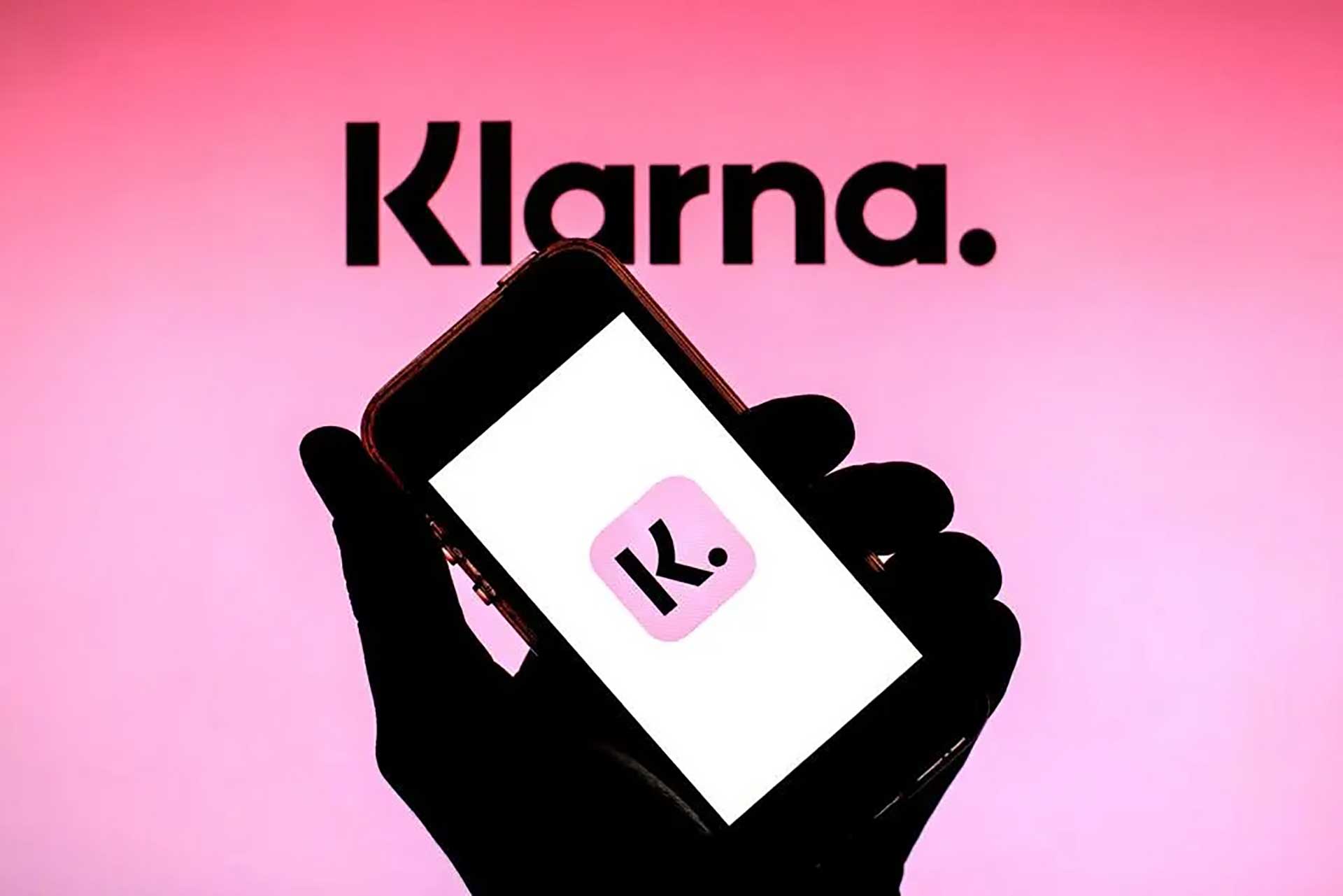 En hand som håller i en mobiltelefon som visar en rosa logotyp med bokstaven "K" och texten "Klarna." ovan, på rosa bakgrund, digital illustration.